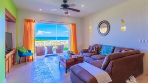 Ein Wohnzimmer mit einer Couch, einem Couchtisch und einem Fernseher. Ein Fenster mit Vorhängen bietet Blick auf den Ozean.