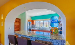 A kitchen with a granite countertop, a water dispenser, and a view of the backyard through a window. - Isla 33 Stunning Oceanfront Villa Bienvenida (La Gloria, Isla Mujeres)