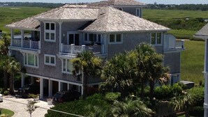 A two-story house with a balcony on the second floor, surrounded by palm trees and a grassy field. - Luxury Coastal Oasis ~ Panoramic Water Views ~ Wrightsville Beach (Wrightsville Beach)