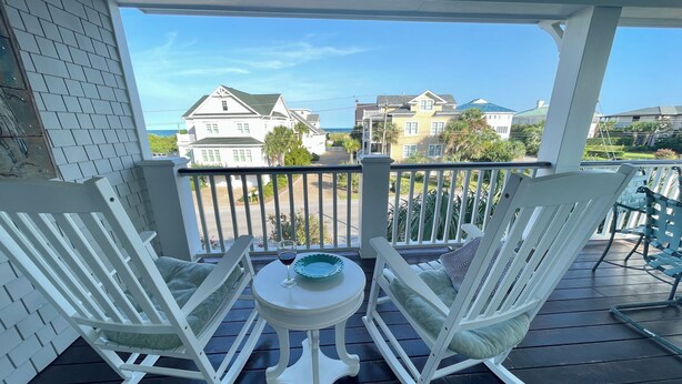 A covered porch with two rocking chairs and a small table, overlooking a residential street. - Luxury Coastal Oasis ~ Panoramic Water Views ~ Wrightsville Beach (Wrightsville Beach)
