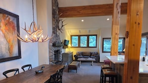 A dining area with a wooden table and chairs, a fireplace, a stone wall, and a sofa in a rustic interior. - Sage Haven Welcomes EIGHT to TEN Guests. Let the Sun Valley Adventures Begin! (Ketchum)