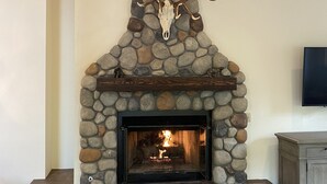 A stone fireplace with a deer skull and antlers above it, a television mounted on the wall, and a leather bench in front. - Sage Haven Welcomes EIGHT to TEN Guests. Let the Sun Valley Adventures Begin! (Ketchum)