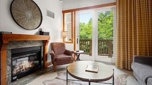 A cozy living room with a fireplace, a leather armchair, a round coffee table, and a view of a balcony with a wooden railing. - 1117 | Studio | The Mansfield | 1st floor | Private Wooded Mountain Views (Stowe)