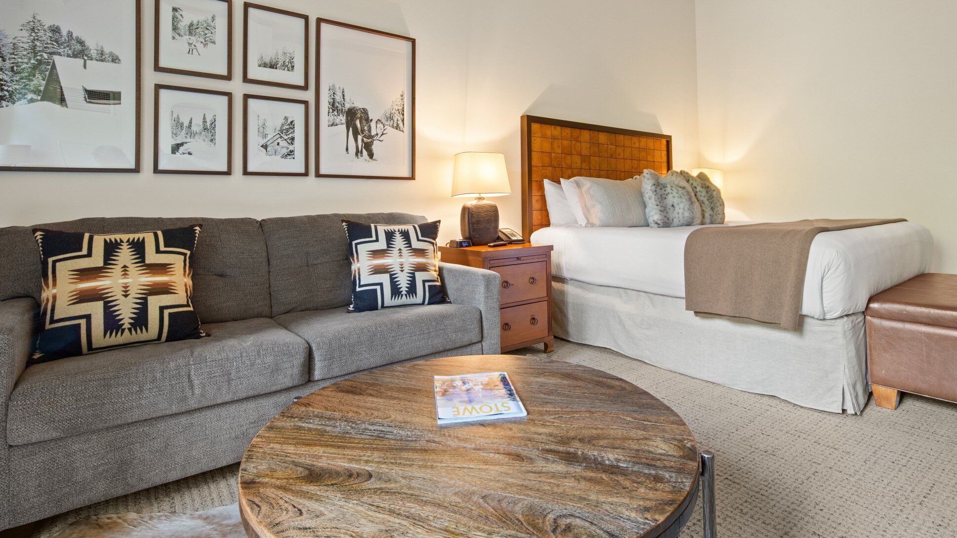Una habitación de hotel acogedora con un sofá gris, una mesa de centro de madera, una cama con cabecero y fotografías enmarcadas en la pared.