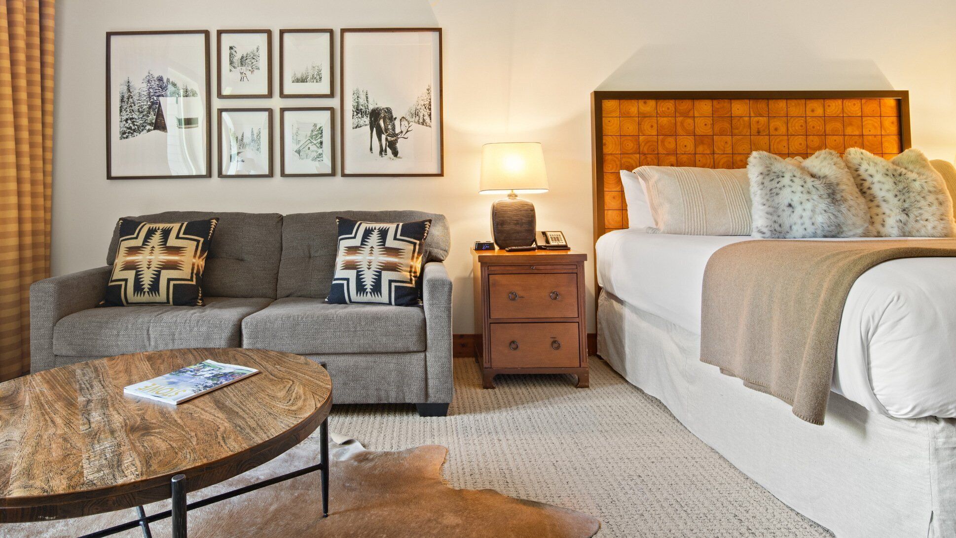 A hotel room with a sofa, a round wooden table, a bed with a headboard, a nightstand with a lamp, and framed pictures on the wall.