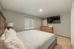 Un dormitorio con cama, cómoda, ventana y televisión de pantalla plana montada en la pared.
