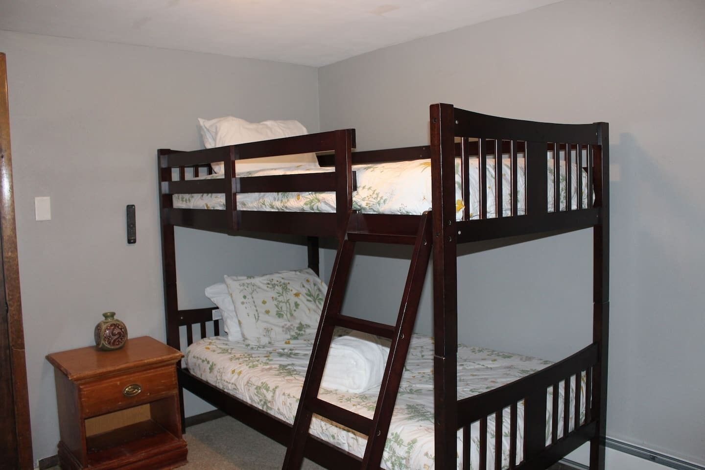 A bunk bed with a ladder, a bedside table with a vase, and a wall switch.