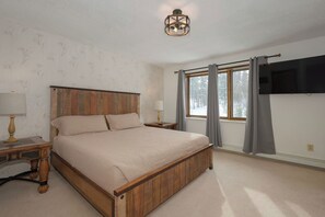 Quarto com cama de madeira, duas mesas de cabeceira com luminárias, janela com cortinas e televisão na parede.