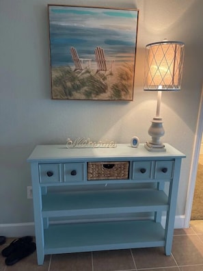 Une console bleu clair, ornée d’une lampe décorative, d’un tableau encadré représentant des chaises de plage et d’une pancarte portant l’inscription « Souvenirs ».