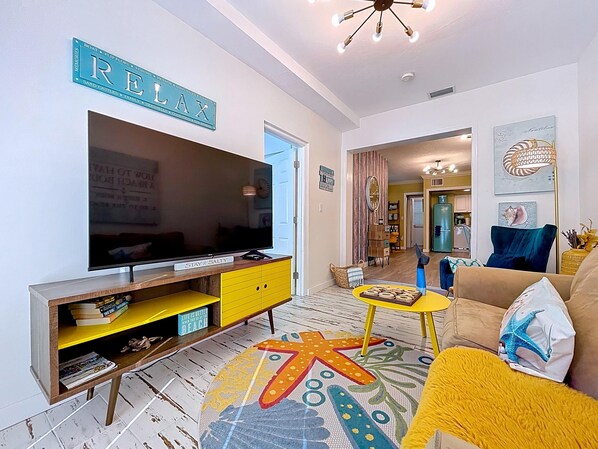 Una sala de estar con un televisor de pantalla plana grande, un sofá amarillo, un tapete con estampado de estrella de mar y un cartel con la palabra 'Relax'.