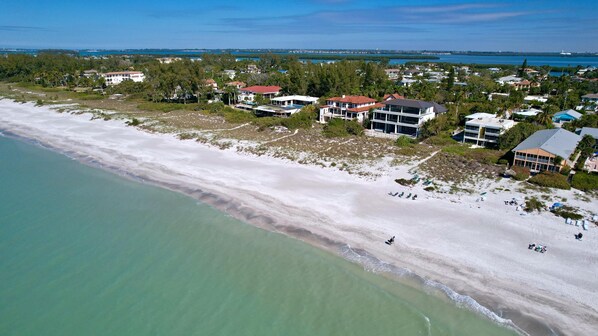 An aerial view of a coastal area with a sandy beach, clear turquoise waters, and several buildings with multiple stories and balconies.