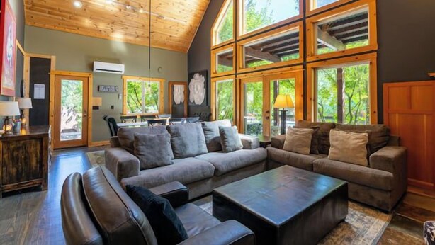 A spacious living room with a large sectional sofa, a coffee table, and a fireplace. There are large windows providing natural light and views of the outdoors.