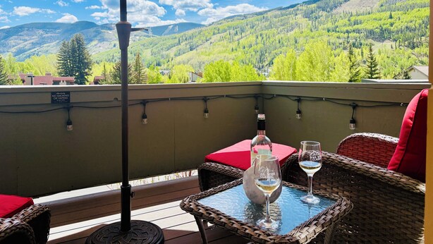 A balcony with wicker furniture, a table with a bottle and glasses, and a view of green mountains.