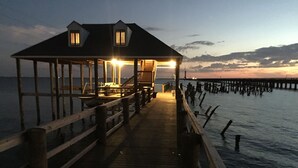 A wooden pier extending into calm waters at dusk, leading to a building with a covered area and two visible windows. - LAKE House with Private Heated Pool, Great for Family Gatherings. Best View! (Slidell)