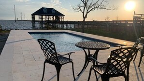A swimming pool with two chairs and a table, a wooden deck, and a view of the water and a distant pier. - LAKE House with Private Heated Pool, Great for Family Gatherings. Best View! (Slidell)
