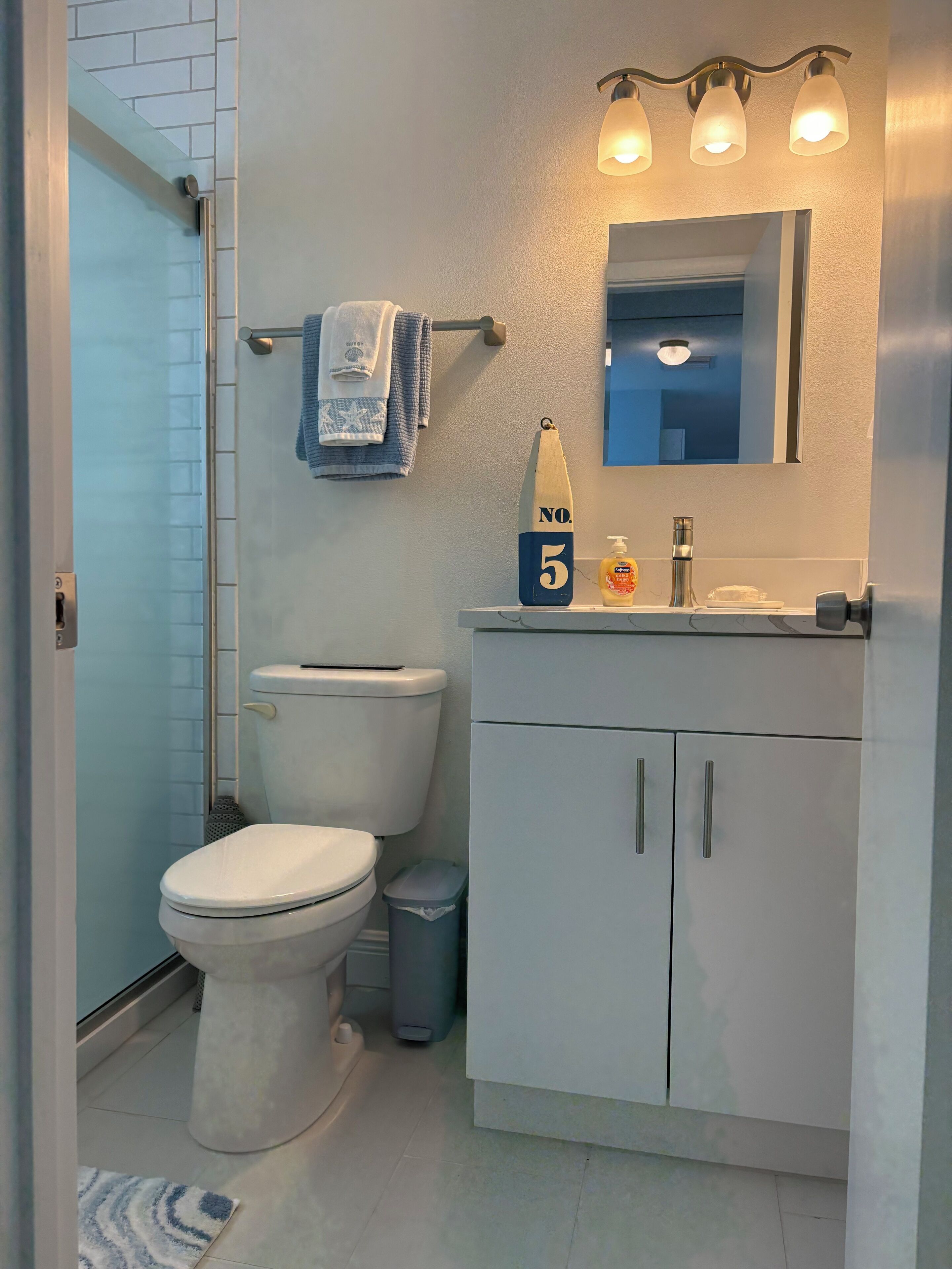 Un baño con inodoro, lavamanos con espejo, toallero y cubo de basura.