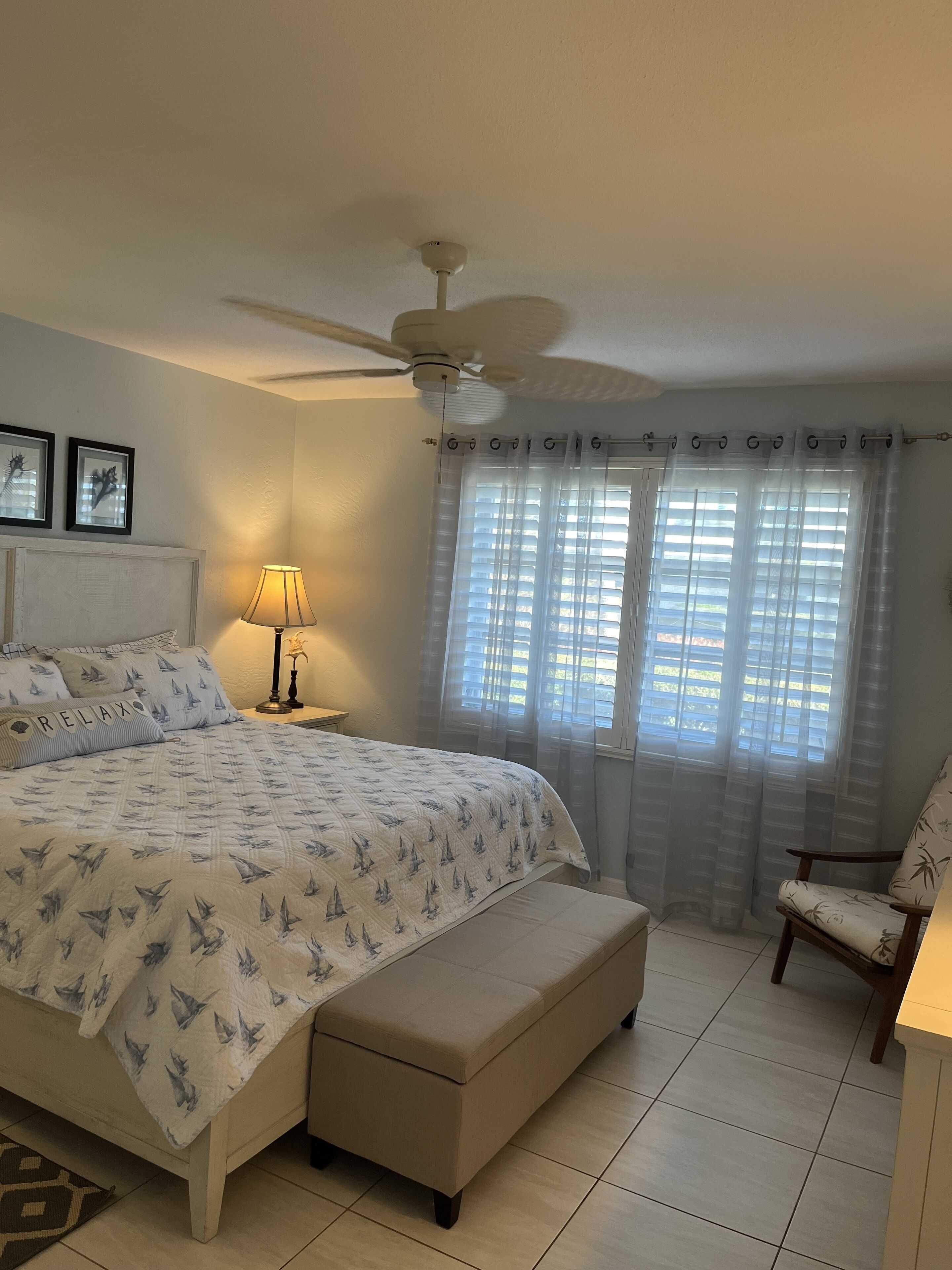 A bedroom with a bed, a ceiling fan, a window with sheer curtains, a bedside lamp, and a chair.