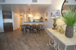 A dining area with a wooden table and chairs, a kitchen in the background, and a decorative lamp on a console table.