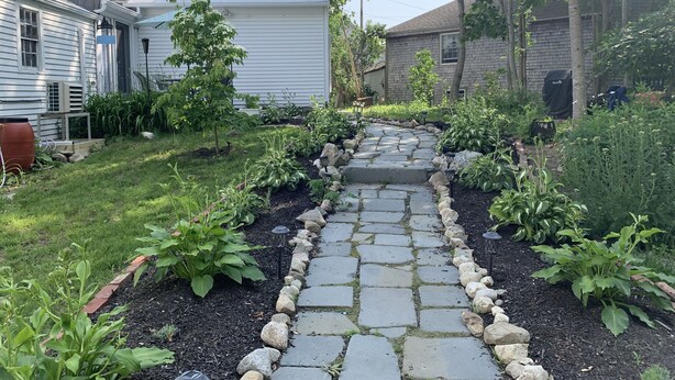 A garden pathway leading to a house with a brick chimney, surrounded by various plants and a small pond. - Discover Coastal charm, just a quick 26-min drive away (Damariscotta)