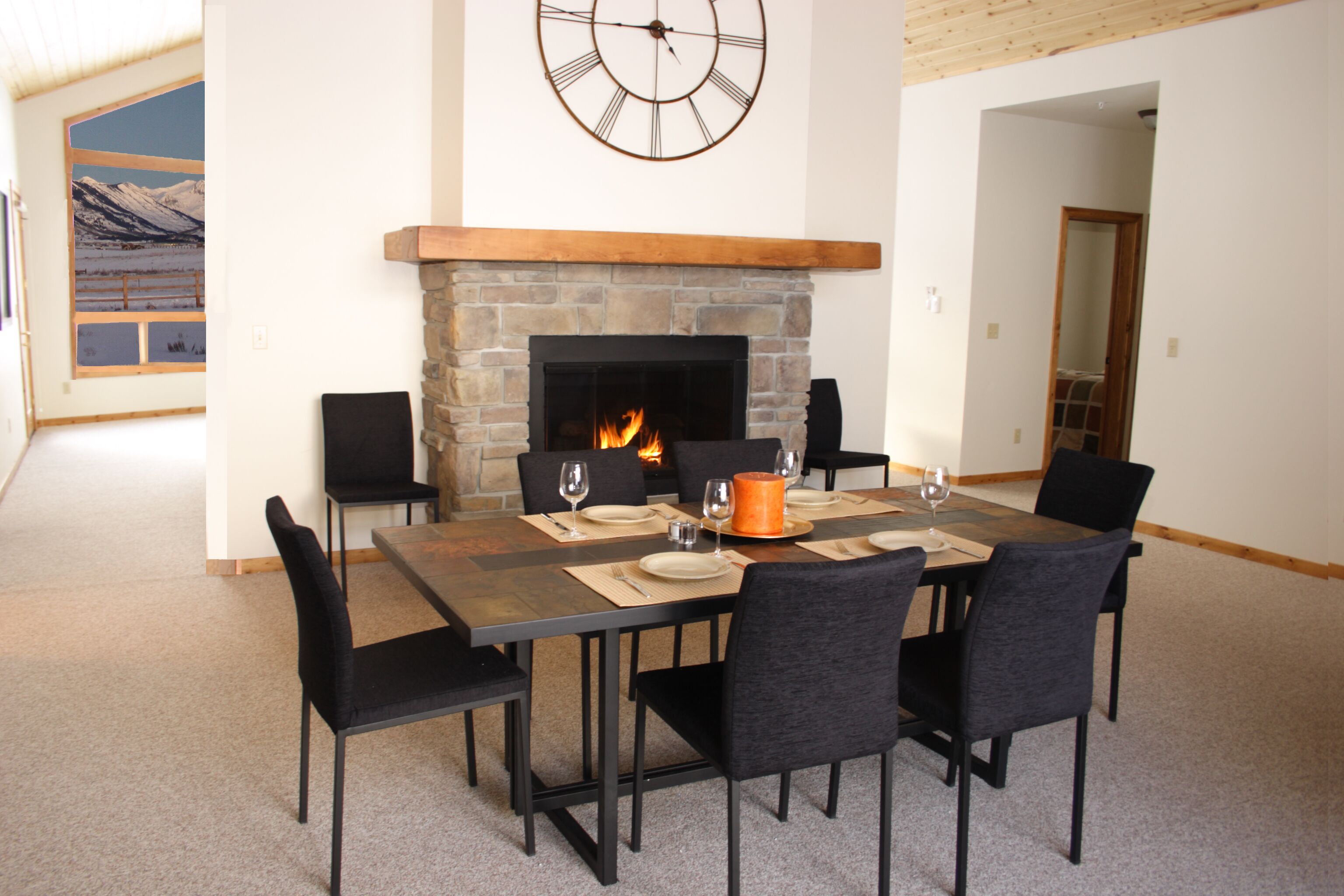 Uma sala de jantar com lareira de pedra, um relógio e vista para montanhas pela janela.