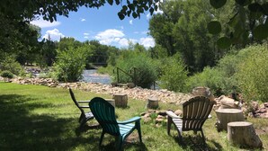 A riverside seating area with two Adirondack chairs, a wooden bench, and a rustic wooden table.