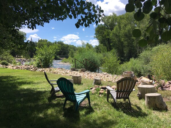 A riverside seating area with two Adirondack chairs, a wooden bench, and a rustic wooden table.