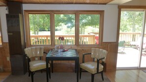A dining area with a wooden table and four chairs, large windows with a view of a deck and trees, and a wooden ceiling.