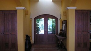 Un pasillo con un portón doble que da a vista de jardín, flanqueado por puertas de madera y un piso de baldosas.