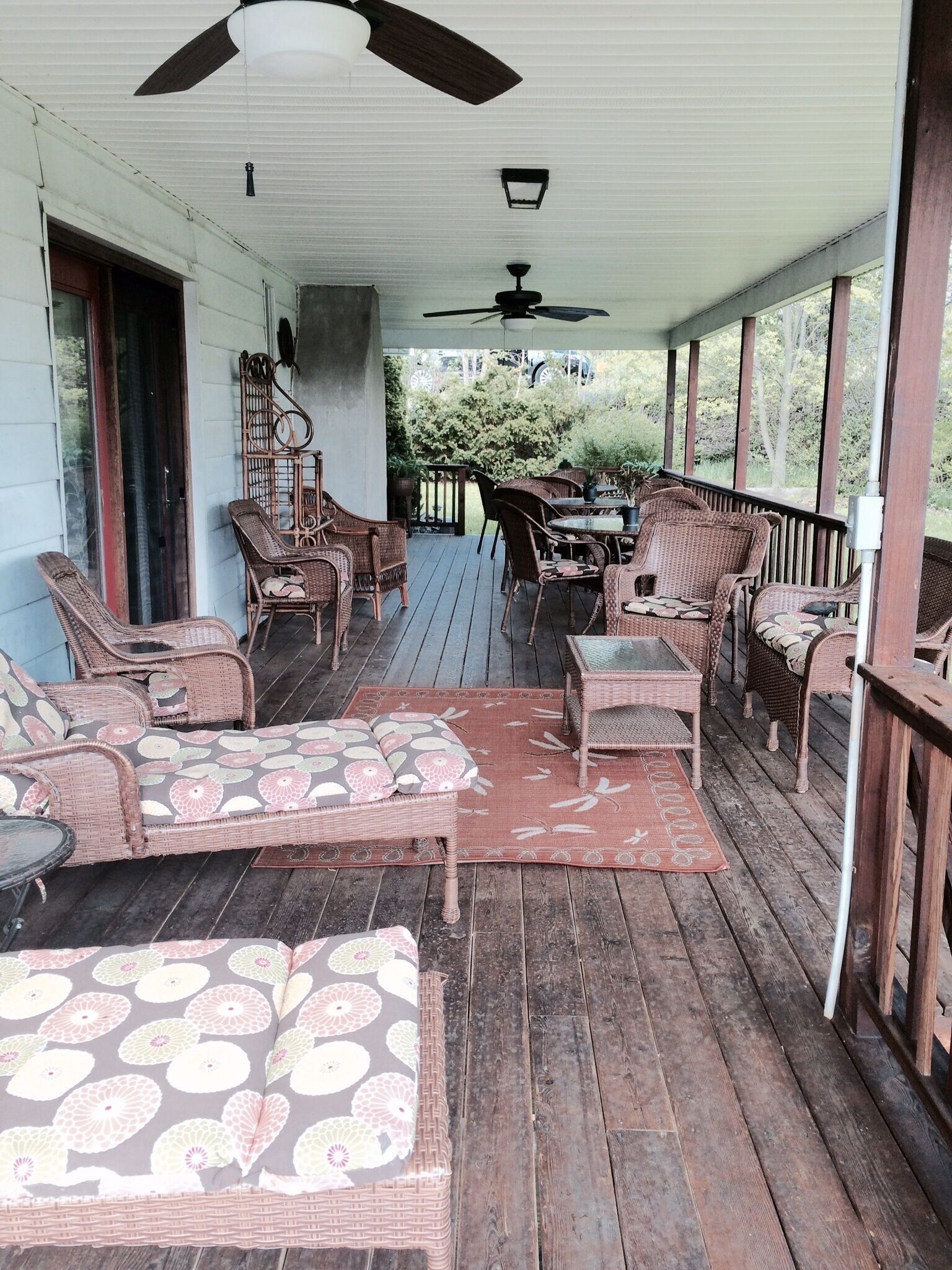 Une véranda couverte, aménagée avec des meubles en osier et une table, dotée d’un ventilateur de plafond et offrant une vue sur la verdure.