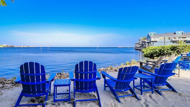 A beachside view with blue Adirondack chairs and a table, overlooking a calm sea and a distant shoreline with buildings. - Water front, Bring Your Boat or drop a fishing line, enjoy a beautiful sunset ! (Punta Gorda)