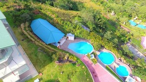 Vue aérienne d’une propriété dotée de plusieurs piscines, d’un grand espace couvert et d’une végétation luxuriante.