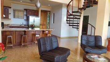 A modern kitchen with wooden cabinets, a stainless steel refrigerator, and a bar area. There are two leather chairs and a glass coffee table in the living area. A spiral staircase is visible in the background.