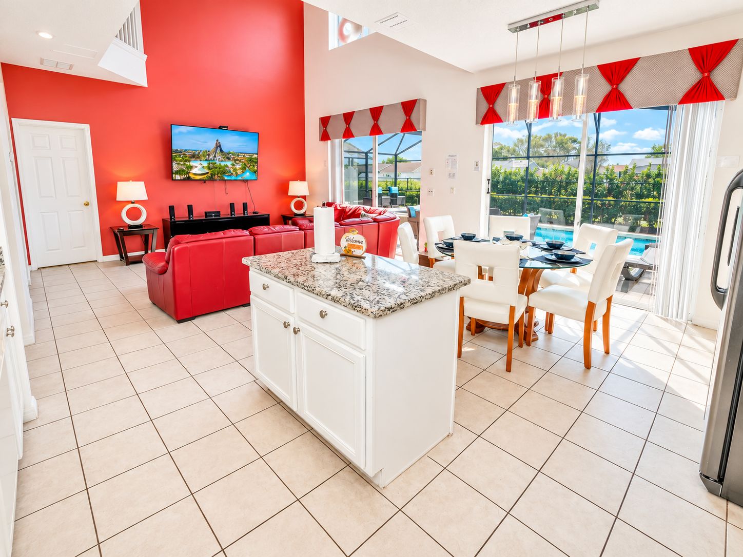A modern kitchen with a central island, a dining area with white chairs, a red sofa, and a large window with a view of greenery.