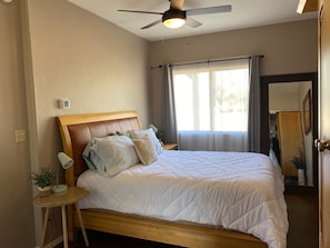 A bedroom with a wooden bed, a ceiling fan, a bedside table with a lamp, and a window with curtains.