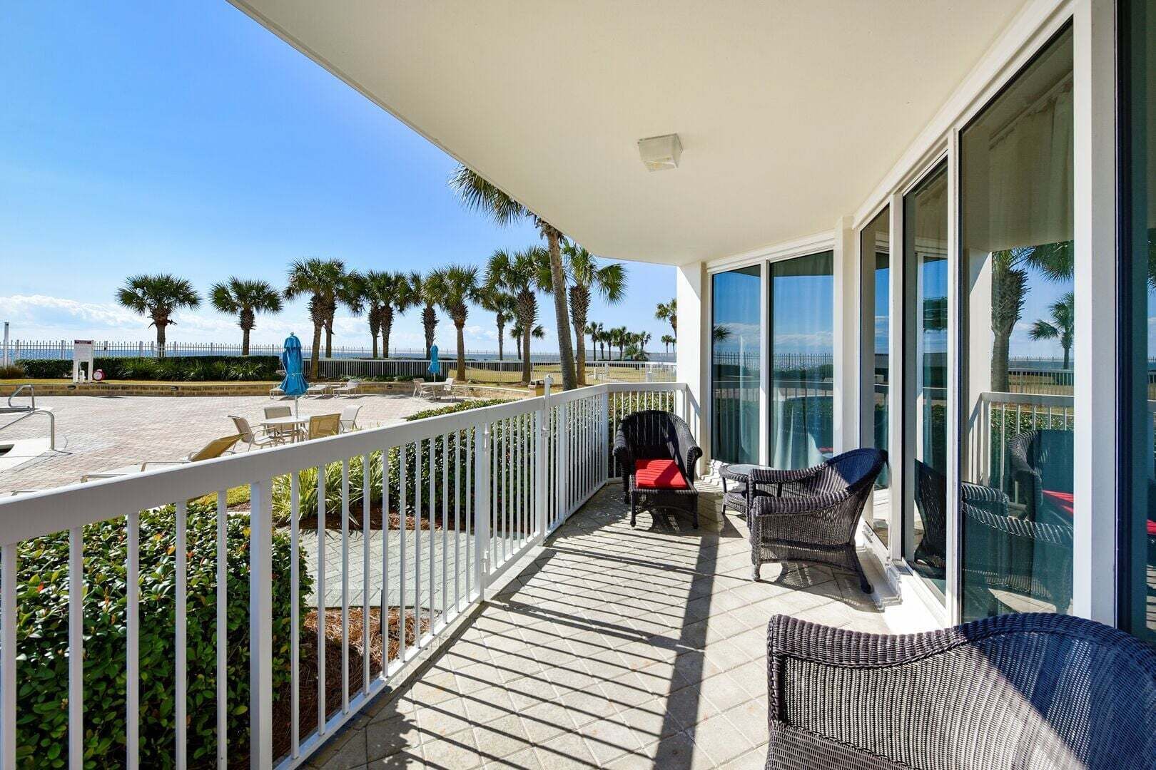 A balcony with wicker furniture, a white railing, and a view of the beach and palm trees.
