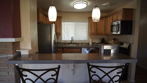 A kitchen with wooden cabinets, a stainless steel refrigerator, a microwave, and a dishwasher. There is a window with blinds and two pendant lights above the island. - Remodeled in-town beauty with Mountain Views! Large, fully fenced back yard. (Kanab)
