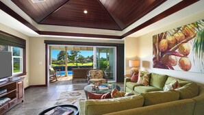 Ein großzügiges Wohnzimmer mit einem großen, grünen Sofa, einem Couchtisch und einem Fernseher auf einem Holzschrank. Ein großes Schiebe-Glaselement führt zu einer Außenpatio mit Pool und Gartenmöbeln.