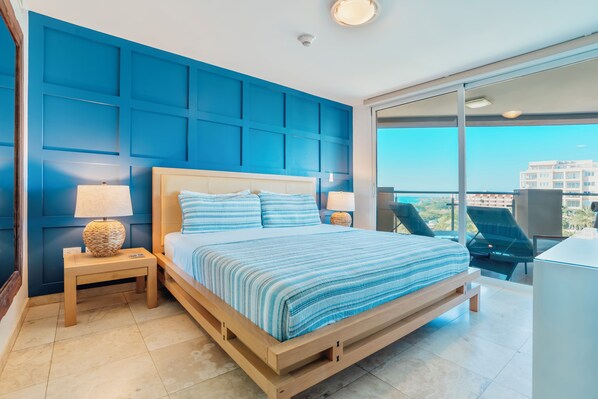 Un dormitorio moderno con una cama grande, dos mesitas de noche con lámparas y vistas a la ciudad a través de amplios ventanales.