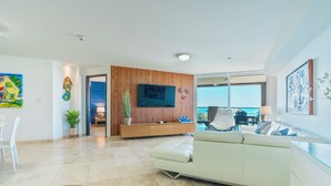Een moderne woonkamer met een witte bank, een wand van houten panelen, een flatscreen televisie en uitzicht op de oceaan door grote ramen.