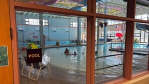 Ein Hallenbad mit Blick durch ein Fenster, ein Liegestuhl und ein Schild, das den Poolbereich kennzeichnet.
