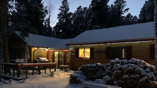 Eine Blockhütte mit einem mit Schnee bedeckten Deck, einer Feuerstelle und einem Holzstapel.