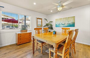 Uma sala de jantar com mesa de madeira, seis cadeiras, ventilador de teto, janela com persianas, uma planta, um quadro e um armário de madeira.