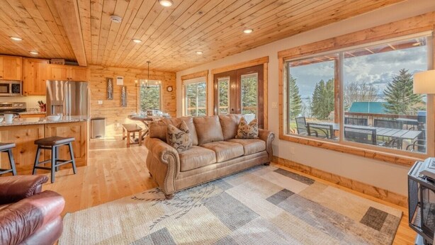 A cozy cabin interior with a wooden ceiling, a leather sofa, a dining area, and large windows offering a view of trees.
