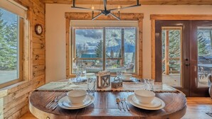 A wooden interior with a dining table set for two, a chandelier, and a large window with a mountain view.