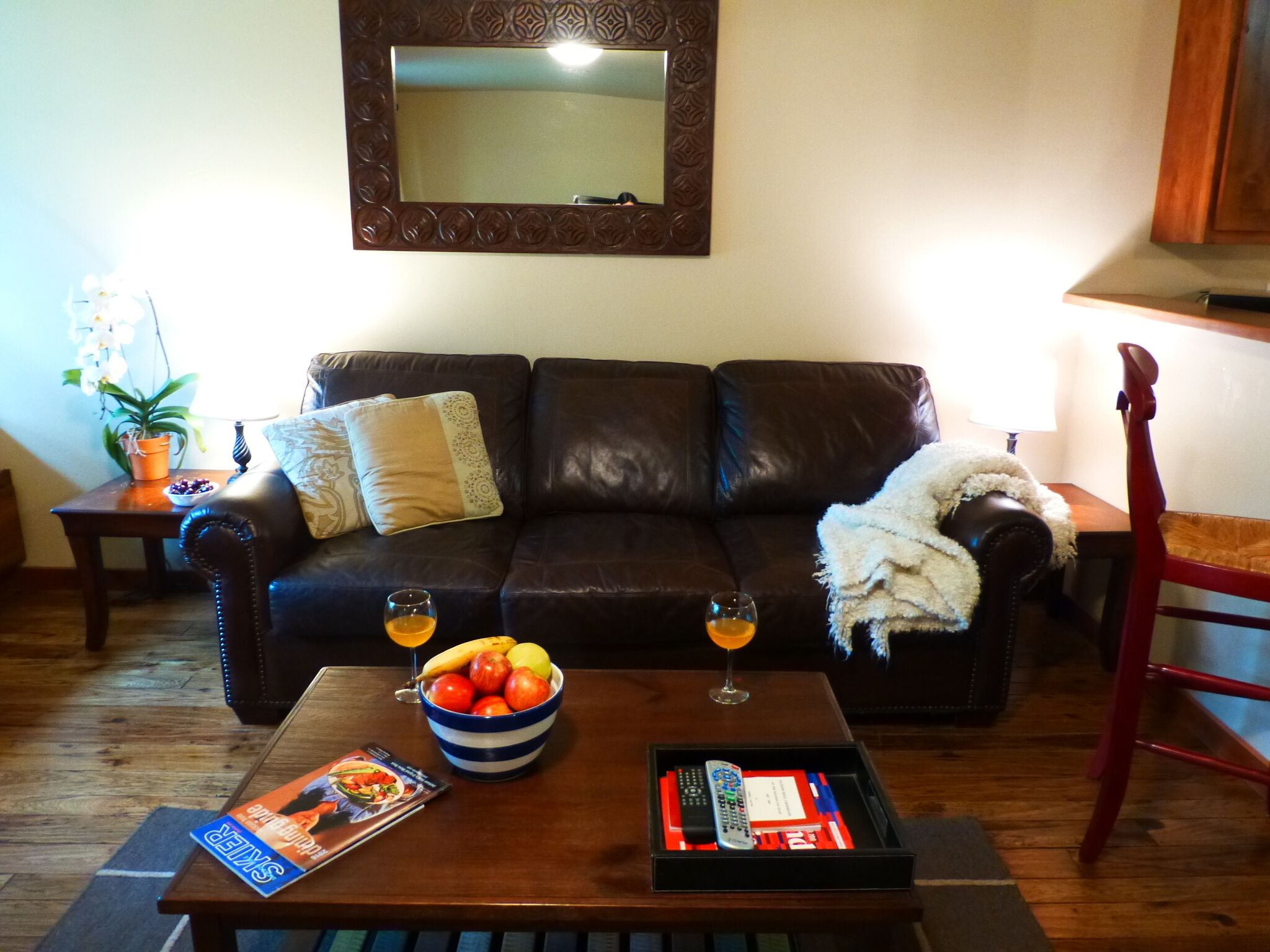 A living room with a dark brown leather sofa, a coffee table with a bowl of fruit and a magazine, a red chair, and a small side table with a lamp.
