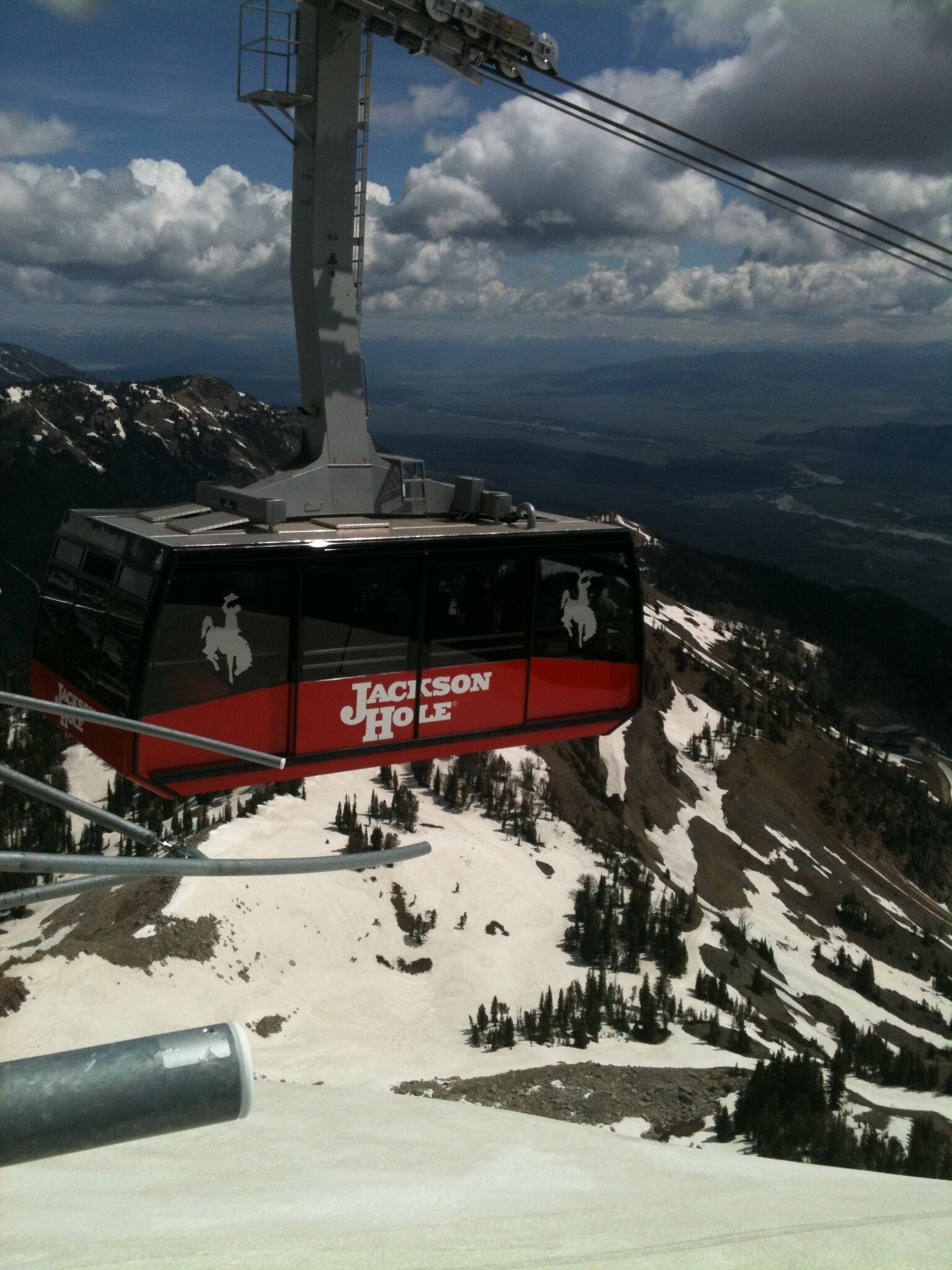 A cable car with "Jackson Hole" branding, suspended over a snowy mountain landscape with trees and distant views.