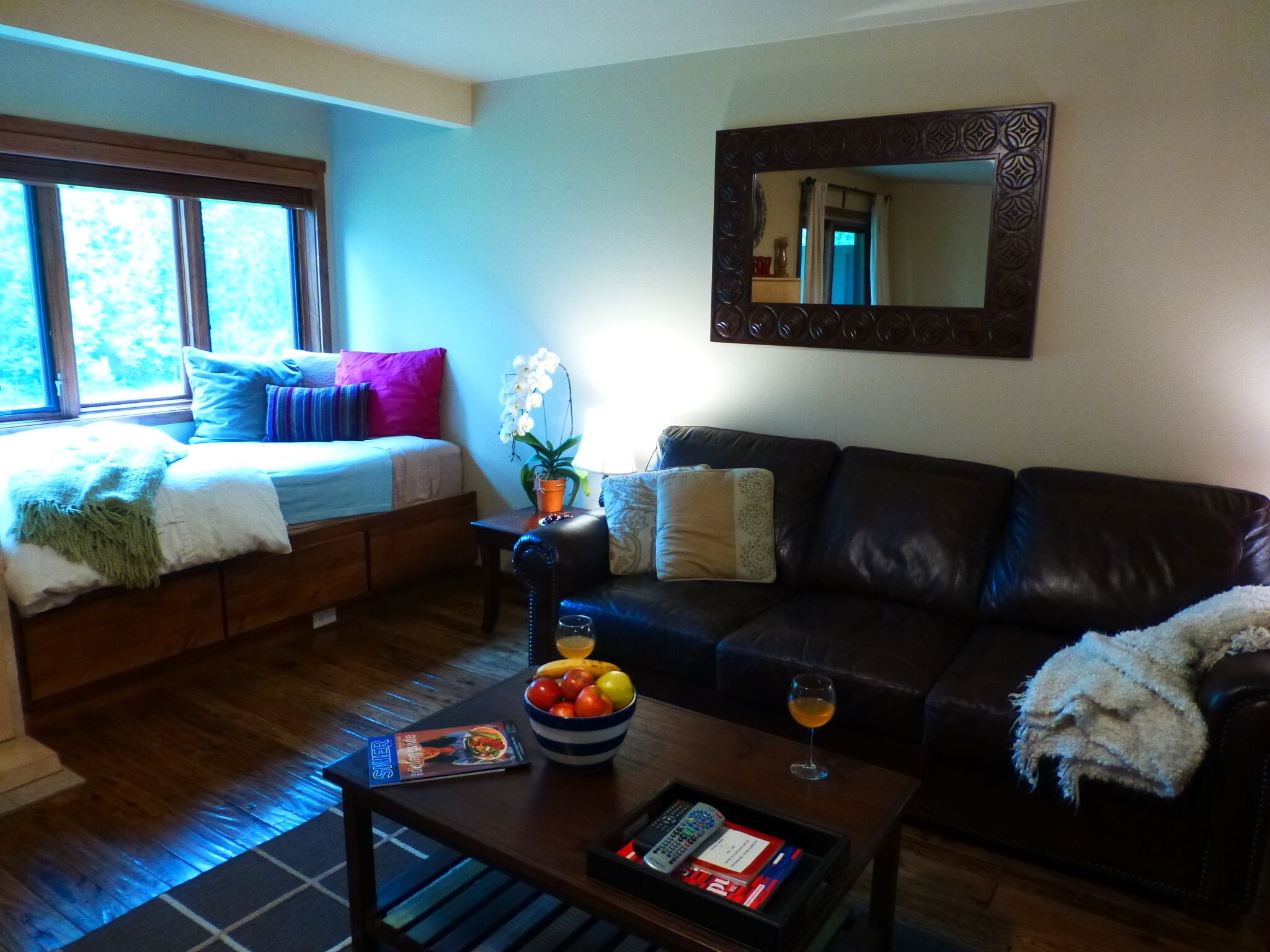Un salon confortable avec un lit, un canapé en cuir, une table basse avec des fruits et une télécommande, et une fenêtre donnant sur des arbres.