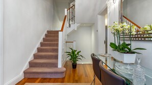Una escalera con peldaños alfombrados que conduce a un segundo piso, un comedor con una mesa y sillas de vidrio, y una planta en maceta sobre el suelo.
