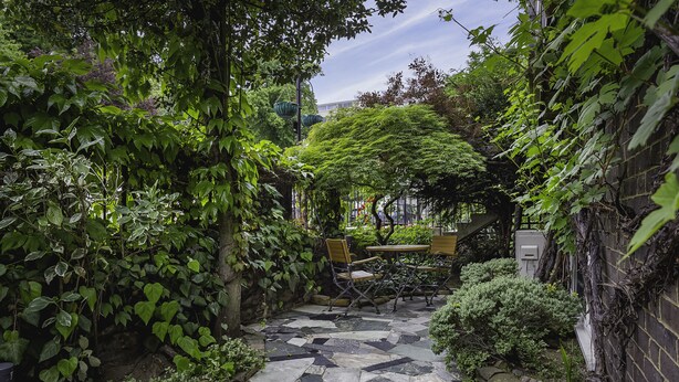 Un sendero de piedra conduce a una mesa y sillas de madera en un exuberante jardín, con abundante follaje y un muro de ladrillo a un lado.