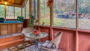 A wooden cabin with a glass-enclosed porch, wicker chairs, a round table with fruit, and a 'Welcome' sign.